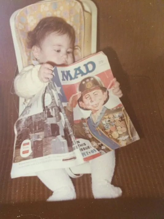 Baby Amanda sitting in a high chair, holding an open copy of MAD magazine as if reading it