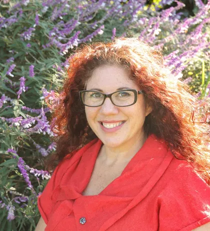 Photo of Amanda Lieber in an orange shirt standing in front of purple flowers.